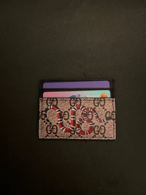 Nuqi korthållare med orm och GG-mönster - Snygg korthållare från G i beige och svart med klassiskt GG-mönster och en röd, vit och svart orm på framsidan. Tillverkad i tåligt canvasmaterial med plats för flera kort. Perfekt accessoar för dig som vill ha något unikt och trendigt.