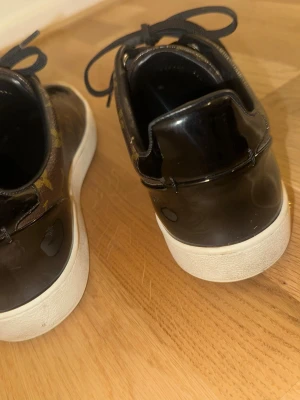 Louis Vuitton monogram sneakers - Bruna sneakers från Louis Vuitton med klassiskt monogrammönster i guld. Skorna har svarta skosnören, svart insida och vit sula. Snygg och tidlös design som sticker ut med exklusiv känsla. Perfekta för dig som gillar lyxiga detaljer och streetstyle.