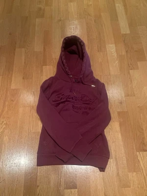 Vinröd hoodie från Superdry - Säljer en vinröd hoodie från Superdry med broderad logga på bröstet och klassisk känguruficka. Hoodien har dragsko i huvan och ribbade muddar vid ärmslut och nederkant. Perfekt för en avslappnad och snygg stil.