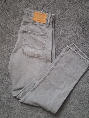 Grå jeans från Jack & Jones - Säljer ett par grå jeans från Jack & Jones med klassisk femficksdesign och rak passform. Jeansen har en snygg vintagegrå tvätt och är tillverkade i slitstarkt denimtyg. Perfekta för dig som gillar en clean och enkel stil.