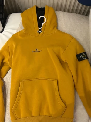 Gul hoodie från Stone Island - Snygg gul hoodie från Stone Island med klassisk logga på vänster ärm och broderad logga på bröstet. Hoodien har magficka framtill och ribbade muddar. Perfekt för dig som vill sticka ut med en streetwear-look.