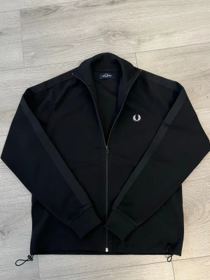Svart ziptröja från Fred Perry - Snygg svart ziptröja från Fred Perry med dragkedja hela vägen, ribbade muddar och diskret logga broderad på bröstet. Tröjan har raka ärmar med svarta band längs axlarna och dragsko i nederkant. Tillverkad i ett mjukt och stretchigt material.