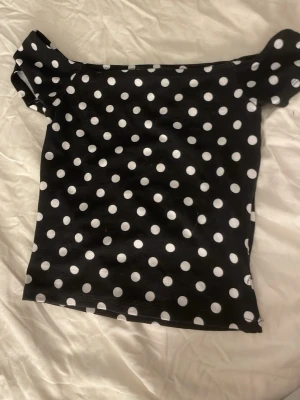 Svart prickig offshoulder topp - Trendig svart offshoulder topp med vita prickar. Snygg passform och korta ärmar som sitter precis vid axlarna. Perfekt för dig som vill sticka ut med ett klassiskt mönster och en ungdomlig vibe.
