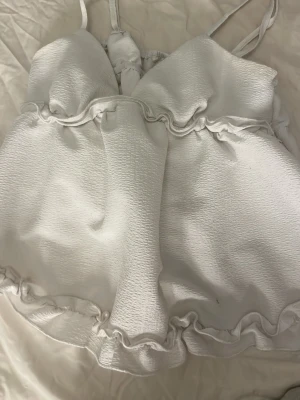 Vit volangtop med tunna axelband - Söt vit topp med tunna axelband och volangdetaljer både upptill och nedtill. Tillverkad i ett lätt, strukturerat tyg som ger en luftig känsla. Perfekt för sommarens alla tillfällen och enkel att matcha med jeans eller kjol.