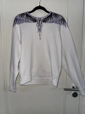 Vit sweatshirt med lila vingmönster - Säljer en vit sweatshirt med ett unikt lila och svart vingmönster över axlar och bröst. Tröjan har rund halsringning, ribbade muddar vid ärmslut och nederkant. Perfekt för dig som vill sticka ut med en cool och konstnärlig design. Materialet känns mjukt och bekvämt.