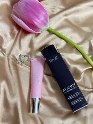 Dior Addict Lip Glow Butter - Lyxigt läppbalsam från Dior i serien Addict Lip Glow Butter. Tuben är ljusrosa med silverfärgat lock och har en elegant metallisk Dior-detalj. Ger återfuktade och mjuka läppar med en subtil glans. Perfekt för dig som vill ha både vård och stil.