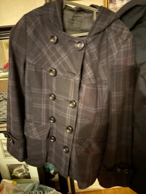 Mörkblå rutig rock med huva - Snygg mörkblå rock med rutigt mönster och dubbelknäppning framtill. Rocken har en huva, stora fickor och dekorativa knappar vid ärmsluten. Passar perfekt för dig som gillar en klassisk men ändå ungdomlig stil.