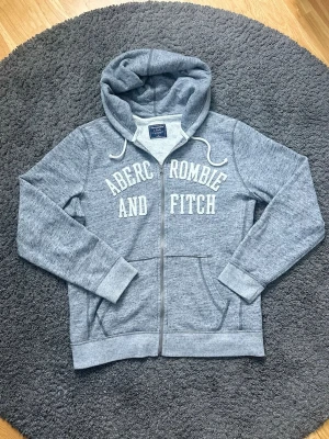 Hoodie från Abercrombie & Fitch - Snygg grå zip-hoodie från Abercrombie & Fitch med vit broderad logga på bröstet. Hoodien har dragsko i huvan, två fickor fram och mjukt material i bomullsblandning. Det är en mer vintage modell, som är lite sällsyntare. 