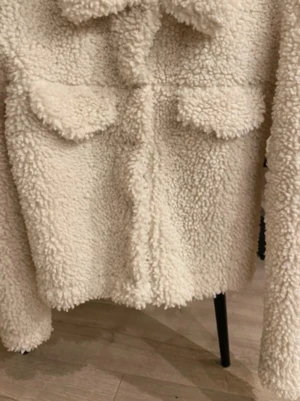 Krämvit teddyjacka med fickor - Mysig krämvit teddyjacka i fluffigt material med två bröstfickor och krage. Jackan har en rak passform och stängs framtill. Perfekt för dig som vill ha en trendig och varm look under kalla dagar.