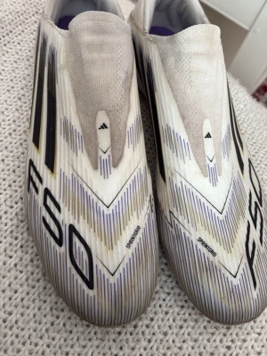 Adidas vita F50 fotbollsskor - Säljer ett par Adidas F50 fotbollsskor i vitt med svarta och gula detaljer. Skorna har en strömlinjeformad design utan snörning och är tillverkade i syntetmaterial för lätt vikt och snabbhet. Perfekta för dig som vill ha speed på planen.