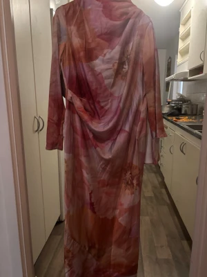 Lång blommig klänning med drapering - Säljer en elegant långklänning med stora blommiga mönster i rosa, beige och orange toner. Klänningen har en unik draperad design, lång ärm på ena sidan och ärmlös på andra, vilket ger en cool oneshoulder-look. Materialet känns glansigt och mjukt, troligen polyester eller satin.