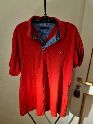 Röd pikétröja från Tommy Hilfiger - Röd pikétröja med kort ärm från Tommy Hilfiger. Klassisk krage med mönstrad insida i blått och vitt, samt liten logga på bröstet. Perfekt för en avslappnad och stilren look.