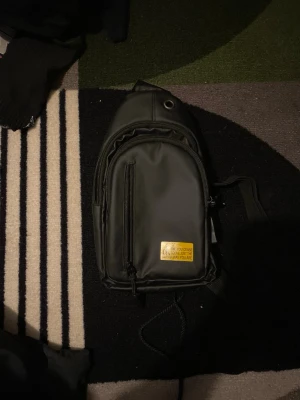 Svart ryggsäck med gul patch - Snygg svart ryggsäck från Calvin Klein med gul patch framtill där det står 'ck and you cease to live just the way you are'. Ryggsäcken har en smidig framficka med dragkedja och en enkel, modern design. Perfekt för dig som vill ha en stilren och praktisk väska.