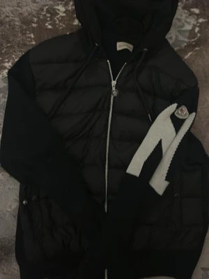 Svart dunväst med huva från Moncler - Snygg svart dunväst från Moncler med huva och dragkedja framtill. Västen har två fickor med knappstängning och ett stort vitt M på ena sidan. Klassisk Moncler-logga på bröstet och cool streetwear-vibe.