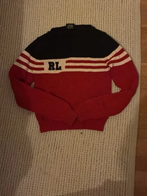 Röd och marinblå stickad tröja RL - Stickad tröja i rött och marinblått från Ralph Lauren med vita och röda ränder över bröstet och RL-bokstäver broderade på vänster sida. Tröjan har långa ärmar, ribbade muddar och en klassisk, sportig look. Sitter som XS
