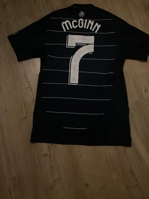 Aston Villa svart matchtröja McGinn - Snygg svart Aston Villa matchtröja från Adidas med vita ränder och tryck. Nummer 7 och McGinn på ryggen, klubbmärke och Betano-logga på bröstet samt vita Adidas-ränder på axlarna. Perfekt för dig som älskar fotboll och vill ha en stilren supportertröja. Köpt på villa park