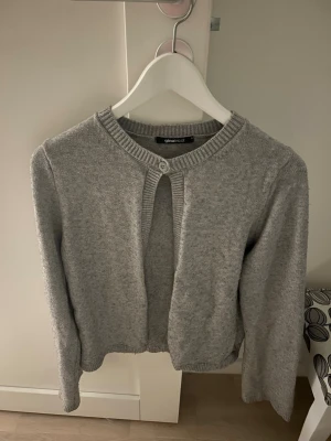 Grå kofta från Gina Tricot - Mysig grå kofta från Gina Tricot med rund halsringning och ribbade kanter. Koftan har en knapp upptill och är öppen framtill, vilket ger en avslappnad look. Perfekt att slänga över en topp för extra lager.