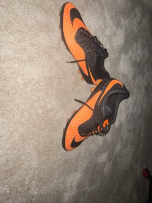 Nike orangea och svarta fotbollsskor - Säljer ett par grymma fotbollsskor från Nike i orange och svart. Skorna har klassisk snörning, mönstrad ovandel för bättre bollkontroll och dobbar för spel på gräs. Perfekta för dig som vill sticka ut på planen och få bra grepp i spelet.