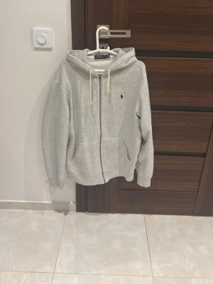 Grå zip-hoodie från Polo Ralph Lauren - Snygg grå hoodie med dragkedja från Polo Ralph Lauren. Klassisk design med huva och vita snören, två fickor framtill och liten broderad logga på bröstet. Perfekt för en avslappnad och stilren look.