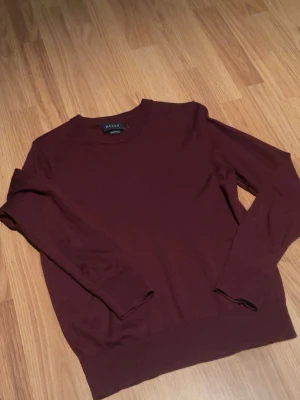 Vinröd stickad tröja från Riley - Mysig vinröd stickad tröja från Riley med rund halsringning och långa ärmar. Tröjan har en enkel, stilren design och ribbade muddar vid ärmslut och nederkant. Perfekt för lager på lager och passar till det mesta.