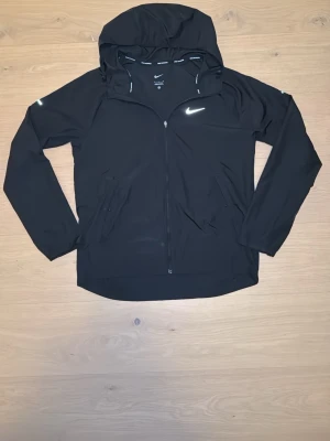 Svart Nike vindjacka med huva - Snygg svart vindjacka från Nike med huva och dragkedja framtill. Jackan har två sidofickor med dragkedja och en vit Nike-logga på bröstet. Tillverkad i lätt och smidigt material, perfekt för blåsiga dagar. Reflekterande detaljer på axlarna för extra synlighet. Vindjackan har dock en liten defekt på baksidan (se sista bilden)