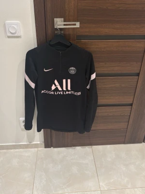 Svart PSG träningströja från Nike - Svart långärmad träningströja från Nike med PSG-logga och rosa detaljer. Halvlång dragkedja framtill, tryck med 'Accor Live Limitless' och 'Visit Rwanda' på ryggen. Perfekt för fotbollsträning eller chill. Materialet är troligtvis polyester.