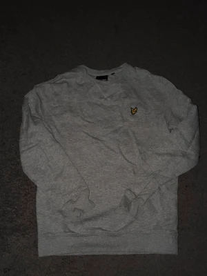Grå sweatshirt från Lyle & Scott - Klassisk grå sweatshirt från Lyle & Scott med rund halsringning och den ikoniska gula örnloggan broderad på bröstet. Tröjan har ribbade muddar vid ärmslut och nederkant. Perfekt för en avslappnad och stilren look. Nypris 950kr