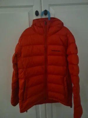Röd dunjacka från Peak Performance - Snygg röd dunjacka från Peak Performance med huva och dragkedja framtill. Jackan har en puffig design med breda kanaler och logga på bröstet samt ärmen. Perfekt för kalla vinterdagar och riktigt varm tack vare dunfyllningen.
