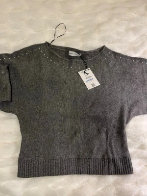 Grå Bershka tröja - Mysig grå stickad tröja från Bershka med pärldetaljer längs axlar och ärmar. Tröjan har ribbstickad nederkant och ärmar, samt en avslappnad passform. Perfekt för att addera lite extra stil till din outfit.