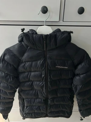Svart dunjacka från Peak Performance - Snygg svart dunjacka från Peak Performance med huva och dragkedja framtill. Jackan är quiltad och har två sidofickor med dragkedja. Perfekt för kalla dagar och har en klassisk look. Passar 160-170 ungefär. Det är tre små lappar på baksidan från hål.