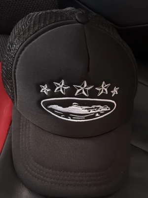 Svart truckerkeps med stjärnor - Snygg svart truckerkeps med mesh på sidorna och broderade vita stjärnor framtill och på sidan. Framtill finns även ett broderat motiv av en bil och fem stjärnor ovanför. Justerbar baktill för perfekt passform.