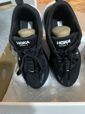 Svarta Hoka Bondi 8 sneakers - En helt ny par Snygga svarta Hoka Bondi 8 sneakers med vit sula och klassisk logga på plösen. Ovandelen är i mesh för bra andningsförmåga och komfort. Skorna har en chunky sula som ger grym dämpning och passar perfekt för dig som gillar streetwear och sportig stil.