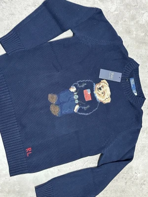 Mörkblå stickad tröja Polo Bear - Mörkblå stickad tröja från Polo Ralph Lauren med den ikoniska Polo Bear-björnen på bröstet. Björnen har jeans, boots och en tröja med amerikanska flaggan. Tröjan har ribbade muddar och RL-broderi nertill. Perfekt för dig som gillar klassisk preppy stil.