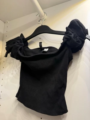 Svart croppad topp med puffärmar - Trendig svart croppad topp med ribbad struktur och stora puffärmar. Toppen har en offshoulder-design och är perfekt för dig som vill sticka ut med en cool och modern stil. Materialet känns mjukt och stretchigt.