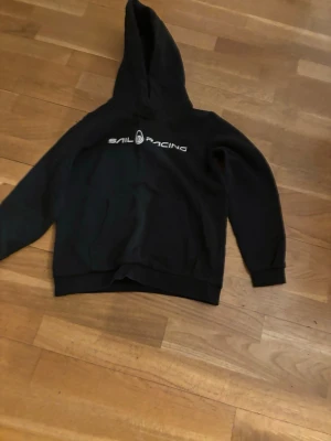 Svart Sail Racing hoodie med tryck - Svart hoodie från Sail Racing med vit logga och text på bröstet. Klassisk modell med huva och känguruficka framtill. Perfekt för en avslappnad och sportig stil. Mjuk bomullsblandning och ribbade muddar vid ärmslut och nederkant.