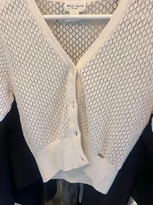 Vit kortstickad kofta Holly & White - Söt vit kort kofta från Holly & White med genombrutet stickat mönster och v-ringning. Koftan har knappar framtill, ribbade muddar och är perfekt att slänga över en topp. Materialet känns mjukt och luftigt, perfekt för vår och sommar.