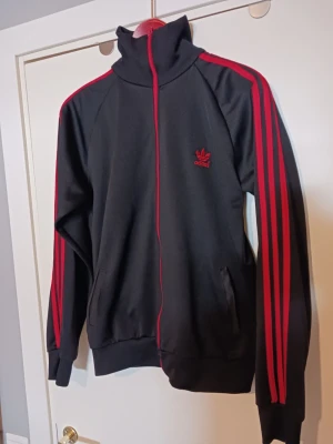 Svart Adidas track jacket med röda ränder Storlek L - Säljer en svart Adidas track jacket med klassiska röda ränder längs ärmarna och röd dragkedja. Jackan har hög krage, två fickor fram och Adidas logga broderad på bröstet. Perfekt för en sportig och retro look. Storlek L
