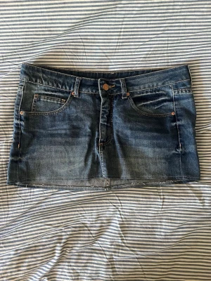 Blå lågmidjad jeanskjol - Snygg mörkblå jeanskjol med klassisk femficksdesign och rå kant nertill. Kjolen har knapp och dragkedja framtill samt en liten läderpatch med logga bak i linningen. Perfekt till alla dina outfits!