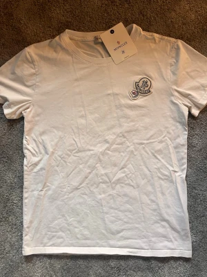 Vit Moncler t-shirt med logga - Säljer en stilren vit t-shirt från Moncler med två broderade loggor på bröstet. Klassisk rund halsringning och korta ärmar. Tillverkad i mjuk bomull som känns skön mot huden. Perfekt för dig som gillar exklusiva och enkla plagg.