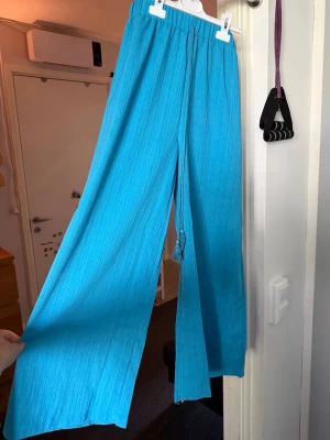 Turkosa vida linnebyxor - Snygga blå byxor med breda ben och resår i midjan. Byxorna har dragsko med tofsar och är tillverkade i ett lätt och luftigt linnematerial. Perfekta för en chill och avslappnad stil.helt nya storlek L