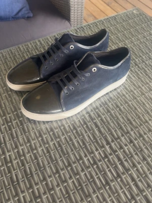 Mörkblå sneakers med lackad tå - Snygga mörkblå lanvins med ovandel i mocka och en glansig, svart lackad tå. Skorna har vita sulor och matchande mörkblå skosnören. Perfekt för dig som gillar en stilren men ändå unik look. Har haft dessa skor under den tidigare sommaren, sitter väldigt bekvämt. De har aldrig blivit blöta så mockan är i suveränt skick. Andledningen att jag säljer de är att jag köpt ett par nya skor samt att de sitter lite trångt på mig.