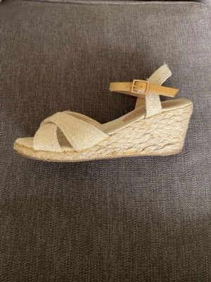Beige kilklackssandal från Next - Snygg beige sandal med kilklack från Next. Ovandelen har korsade band i textil och justerbart ankelband med spänne. Klacken är flätad i repstil för en somrig vibe. Perfekt till jeans eller klänning när du vill ha lite höjd men ändå bekvämt.