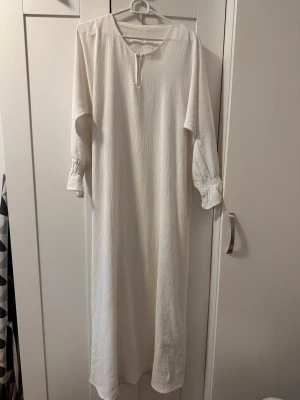 Vit långklänning - En enkel och stilren vit långklänning med rund halsringning och liten öppning framtill. Klänningen har långa ärmar med resår vid ärmsluten för en avslappnad look. Tillverkad i ett lätt, strukturerat tyg som ger en mjuk känsla.