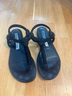 Svarta sandaler från Acne Studios - Stilrena svarta sandaler från Acne Studios med rundad tå och breda mockaband som är knutna framtill. Sandalerna har öppen häl och tå, samt en platt sula för en avslappnad look. Perfekta för sommaren och passar till många olika outfits. Strl 36. Tillverkade i Italien.  Skicket är bra använt – normalt slitage på innersula och yttersula enligt bilderna, men fortfarande mycket fina och fullt användbara.