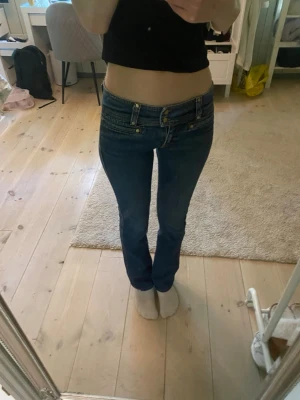 Mörkblå bootcut jeans med låg midja - Säljer ett par mörkblå bootcut jeans med låg midja och dubbla knappar fram. Jeansen har klassiska fickor både fram och bak samt detaljerade sömmar. Det står storlek 40 på dem men de är mer som storlek S. Säljes då de ej kommer till användning