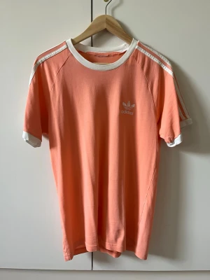 Persikofärgad Adidas Originalt-shirt strl L - Snygg persikofärgad t-shirt från Adidas Original med klassiska ränder på axlarna. T-shirten har korta ärmar, rund halsringning och Adidas-logga på bröstet. Storlek L. Material bomull. Etikett om material och storlek är klippta.