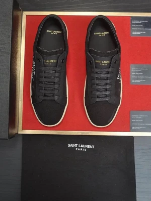 Svarta Saint Laurent sneakers - Snygga svarta sneakers från Saint Laurent med vit sula och handskriven logga på sidan. Skorna har klassisk låg profil, svarta skosnören och detaljer i läder och textil. Perfekta för dig som gillar stilrena och exklusiva sneakers.