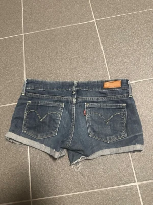 Blå jeansshorts från Levi's low waist - Säljer ett par klassiska blå jeansshorts från Levi's med upprullade benslut och slitna detaljer. Shortsen har fem fickor, bälteshällor och knappgylf. Perfekta för varma dagar och en avslappnad look. W25