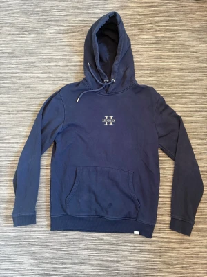 Mörkblå hoodie Les Deux - Mörkblå hoodie från Les Deux med broderad logga på bröstet. Klassisk känguruficka framtill, ribbade muddar och justerbar dragsko i huvan. Tillverkad i mjuk bomullsblandning som känns skön mot huden. Perfekt för en avslappnad och stilren look.