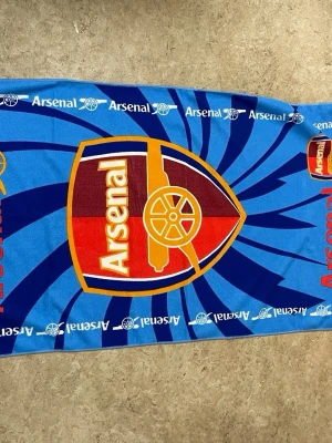 Arsenal badhandduk blå - Säljer en blå badhandduk med stort Arsenal-emblem i rött, gult och vitt. Handduken har ett mönster med klubbens logga och texten 'Arsenal' upprepat längs kanterna. Perfekt för fotbollsfans som vill visa sitt lag på stranden eller gymmet.